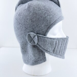 May include: Gray fleece knight helmet with a chin strap and a visor. The helmet is on a white mannequin head.