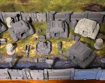May include: A collection of grey 3D printed miniature buildings and structures, including a tall tower, a building with a large door, and a smaller building with a rounded roof. The miniatures are arranged on a textured surface with a green background.