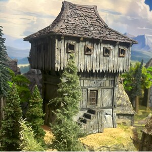 May include: A detailed miniature wooden tower with a weathered, textured appearance. The tower features a steeply pitched, shingled roof, small square windows, and a decorative, notched trim. Green trees and a mountainous backdrop complete the scene.