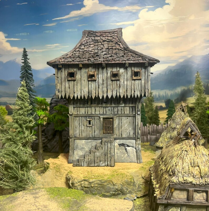 May include: A miniature wooden medieval-style building with a weathered appearance. The structure features a gabled roof, small square windows, and a stone foundation. The scene includes miniature trees and a backdrop of mountains.