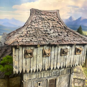May include: A miniature wooden building with a weathered, textured appearance. The roof is made of overlapping wooden shingles. The walls are constructed of vertical wooden planks. Small, square windows with hinged shutters are visible. The background features a landscape with mountains.