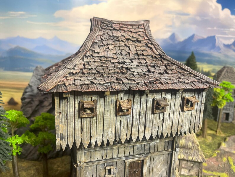 May include: A miniature wooden building with a weathered, textured appearance. The roof is made of overlapping wooden shingles. The walls are constructed of vertical wooden planks. Small, square windows with hinged shutters are visible. The background features a landscape with mountains.
