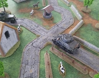 May include: A tabletop wargaming diorama featuring a gray tank and a black military truck on a textured road intersection. The scene includes a small building with a brown roof, a stone well, miniature green trees, stone walls, and small cow figurines.