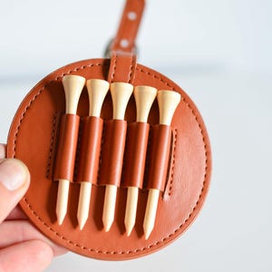 May include: A brown leather golf tee holder with five wooden golf tees. The holder has a stitched edge and a loop for attaching to a belt or bag.