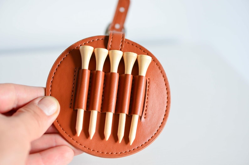 May include: A brown leather golf tee holder with five wooden golf tees. The holder has a stitched edge and a loop for attaching to a belt or bag.