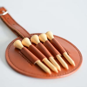 May include: A brown leather golf tee holder with a strap and six wooden golf tees.