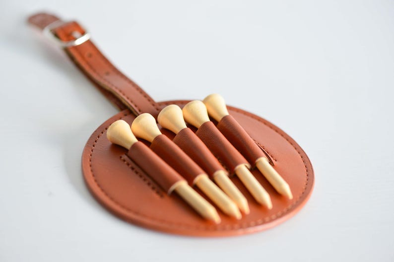 May include: A brown leather golf tee holder with a strap and six wooden golf tees.