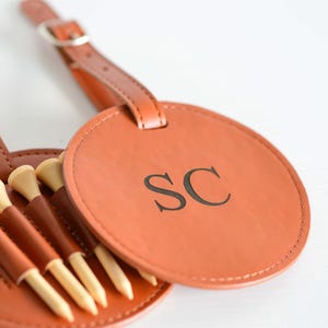 May include: A brown leather golf ball holder with a strap and a circular tag that says "SC". The holder has five slots for golf tees.