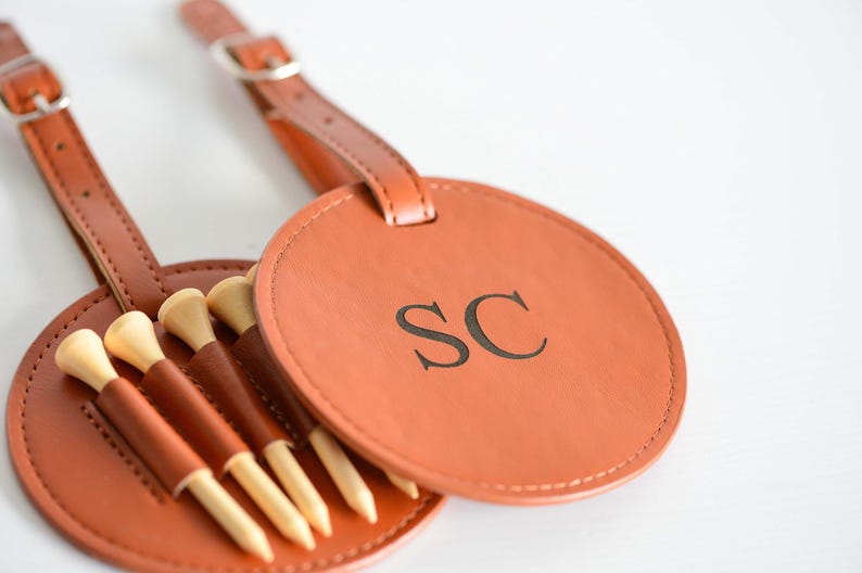 May include: A brown leather golf ball holder with a strap and a circular tag that says "SC". The holder has five slots for golf tees.