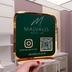 Könnte beinhalten: Ein goldfarbener Rahmen mit grünem Hintergrund und dem Text "Malvales Beauty Lab" in Weiß. Das Schild zeigt auch ein weißes "M"-Logo, ein weißes Instagram-Symbol mit dem Text "@malvales_beauty_lab", einen schwarz-weißen QR-Code und die Telefonnummer "+38 (099) 619 69 69" in Weiß.