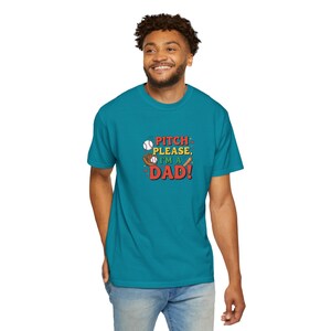 Pode incluir: Uma camiseta azul-turquesa com o texto "PITCH PLEASE, I'M A DAD!" em um design temático de beisebol. O gráfico inclui uma bola de beisebol, uma luva e um bastão. A camisa é de cor sólida e tem gola redonda.