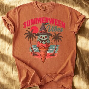 May include: A rust-colored t-shirt featuring a graphic of a Halloween-themed ice cream cone with a witch hat, palm trees, and the text "SUMMERWEEN Vibes". The design includes a red sun and ocean waves.