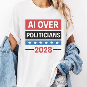 Puede incluir: Camiseta blanca con el texto "AI OVER POLITICIANS - 2028" en rojo, negro y azul. El diseño incluye estrellas y rayas horizontales. La camiseta es de estilo casual.