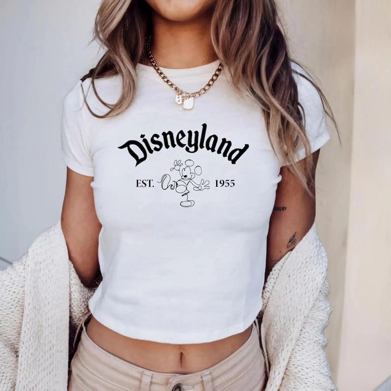 May include: White crop top featuring the Disneyland logo in black script with Mickey Mouse and the text "EST. 1955". The shirt is a casual style, suitable for everyday wear.