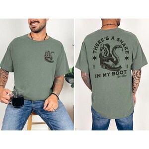 May include: Olive green t-shirt with a graphic of a snake coiled around a cowboy boot. The back of the shirt features the text "THERE'S A SNAKE IN MY BOOT" with a similar graphic. The shirt is made of a soft material.