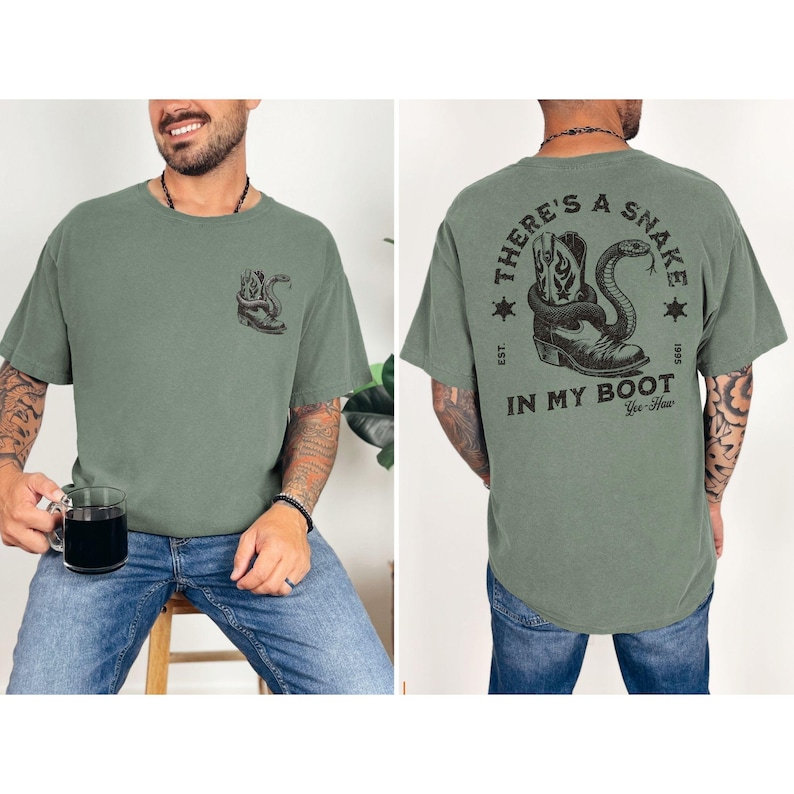 May include: Olive green t-shirt with a graphic of a snake coiled around a cowboy boot. The back of the shirt features the text "THERE'S A SNAKE IN MY BOOT" with a similar graphic. The shirt is made of a soft material.