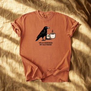 May include: Rust-colored t-shirt featuring a black crow illustration with a coffee cup and the text "Hello Darkness My Old Friend." The shirt has rolled-up sleeves and a relaxed fit, perfect for casual wear.