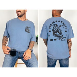 May include: Light blue t-shirt with a black graphic of a snake coiled around a cowboy boot. The text on the back of the shirt reads "THERE'S A SNAKE IN MY BOOT" with additional text and star accents. The shirt is a casual style.