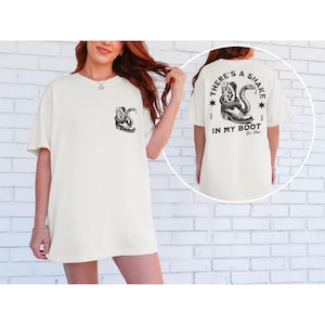 May include: Off-white t-shirt featuring a black graphic of a snake coiled around a cowboy boot on the front and back. The back of the shirt has the text "THERE'S A SNAKE IN MY BOOT" with stars and the year 1995.