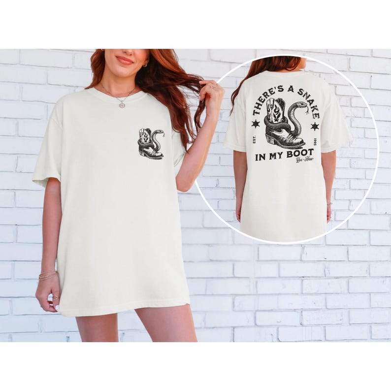 May include: Off-white t-shirt featuring a black graphic of a snake coiled around a cowboy boot on the front and back. The back of the shirt has the text "THERE'S A SNAKE IN MY BOOT" with stars and the year 1995.