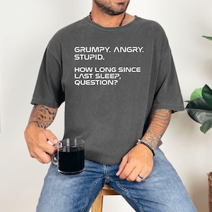 May include: Dark gray t-shirt with white text: "GRUMPY. ANGRY. STUPID. HOW LONG SINCE LAST SLEEP, QUESTION?" The shirt is worn by a person holding a clear glass mug. The person is wearing blue jeans.