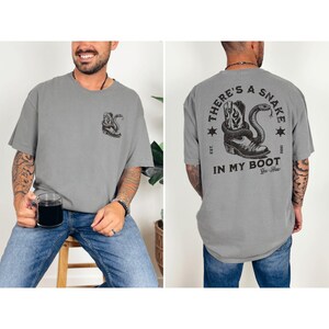 May include: Gray t-shirt with a graphic of a cowboy boot and snake. The back of the shirt features the text "THERE'S A SNAKE IN MY BOOT" in a circular design. The shirt has a relaxed fit.