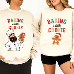 Puede incluir: Sudaderas color crema con el texto "BAKING a little COOKIE" en rojo, verde y naranja. Una sudadera presenta a un chef sosteniendo un hombre de jengibre, la otra un hombre de jengibre con un gorro de Papá Noel. Se ven pantalones cortos y pantalones negros.