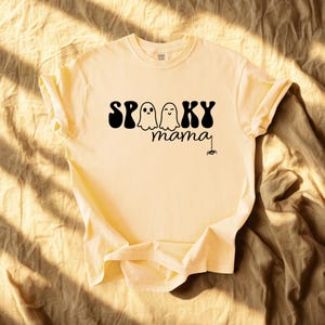 May include: A light yellow t-shirt with the words "Spooky mama" in black, featuring two ghost illustrations and a spider. The shirt has rolled-up sleeves and is made of soft material, perfect for Halloween.