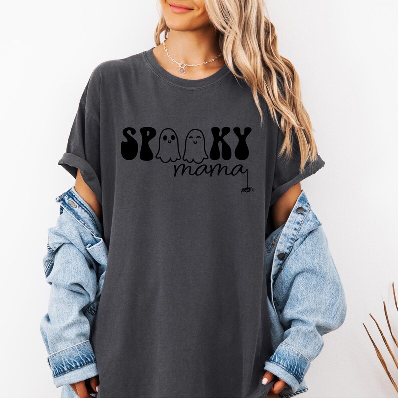 May include: Dark gray t-shirt with the words "Spooky Mama" in black lettering. The word "Spooky" is above the word "mama", with two ghost figures between the words. A small spider is drawn below the word "mama".