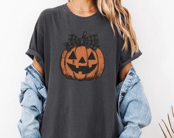 Puede incluir: Camiseta gris oscuro con un diseño de calabaza naranja desgastada y un lazo a cuadros negro y gris. La calabaza tiene una cara tallada. Un estilo cómodo e informal, perfecto para Halloween.