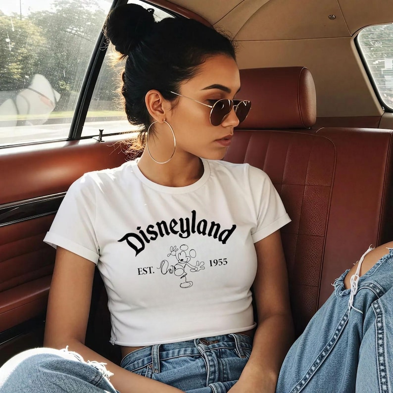 May include: White crop top with the word "Disneyland" in black script, along with an image of Mickey Mouse and the year "1955". The shirt is paired with blue jeans.