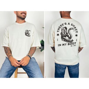 May include: Cream-colored t-shirt featuring a graphic of a cowboy boot and snake. The front of the shirt has a small graphic of the same design. The back of the shirt has the text "THERE'S A SNAKE IN MY BOOT" with additional text and stars.