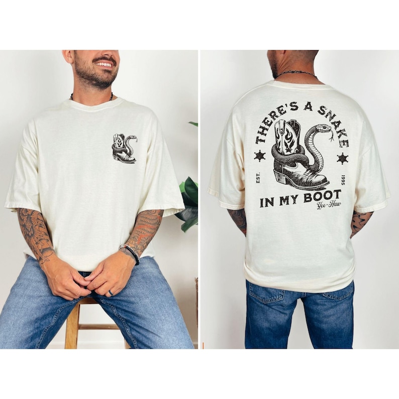 May include: Cream-colored t-shirt featuring a graphic of a cowboy boot and snake. The front of the shirt has a small graphic of the same design. The back of the shirt has the text "THERE'S A SNAKE IN MY BOOT" with additional text and stars.
