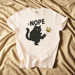 May include: Cream-colored t-shirt with a black cat graphic kicking a gold crown. The word "-NOPE" is above the cat. The shirt has short sleeves and a crew neck.