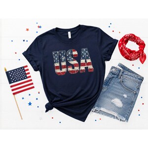 Könnte beinhalten: Ein marineblaues Kurzarm-T-Shirt mit dem Text USA in einem abgenutzten amerikanischen Flaggenmuster. Das T-Shirt wird zusammen mit einer kleinen amerikanischen Flagge an einem Holzstab, einem roten Bandana und hellblauen Jeansshorts gezeigt.