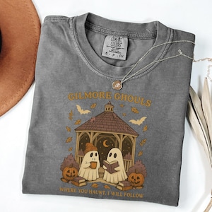 Może przedstawiać: Szara koszulka Comfort Colors z napisem "Gilmore Ghouls" i grafiką przedstawiającą dwa duchy w altanie, dynie i nietoperze. Tekst "Where you haunt, I will follow" znajduje się poniżej grafiki. Srebrny naszyjnik jest zarzucony na koszulkę.