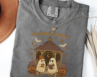 Puede incluir: Una camiseta gris Comfort Colors con el texto "Gilmore Ghouls" y un gráfico de dos fantasmas en una glorieta, calabazas y murciélagos. El texto "Where you haunt, I will follow" está debajo. Un collar plateado está sobre la camiseta.