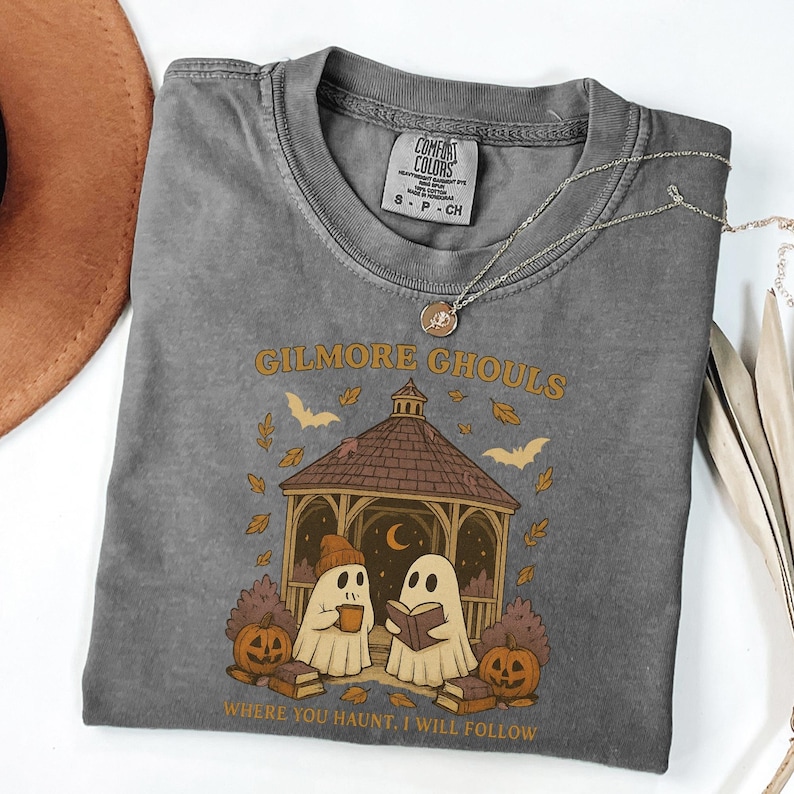Może przedstawiać: Szara koszulka Comfort Colors z napisem "Gilmore Ghouls" i grafiką przedstawiającą dwa duchy w altanie, dynie i nietoperze. Tekst "Where you haunt, I will follow" znajduje się poniżej grafiki. Srebrny naszyjnik jest zarzucony na koszulkę.