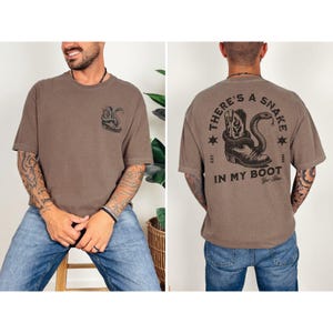 May include: Brown t-shirt with a snake and cowboy boot graphic. The front of the shirt features a snake design, while the back displays the text "THERE'S A SNAKE IN MY BOOT" with a boot and snake illustration. The shirt has short sleeves.
