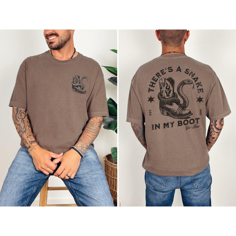 May include: Brown t-shirt with a snake and cowboy boot graphic. The front of the shirt features a snake design, while the back displays the text "THERE'S A SNAKE IN MY BOOT" with a boot and snake illustration. The shirt has short sleeves.