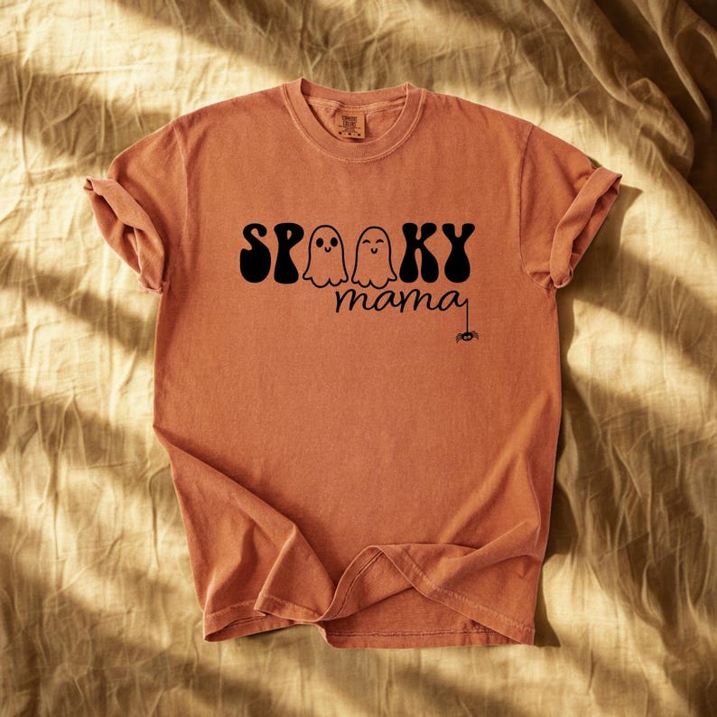 May include: A terracotta-colored t-shirt with rolled-up sleeves. The shirt features the words "Spooky mama" in black, with two ghost illustrations and a spider graphic, perfect for Halloween.