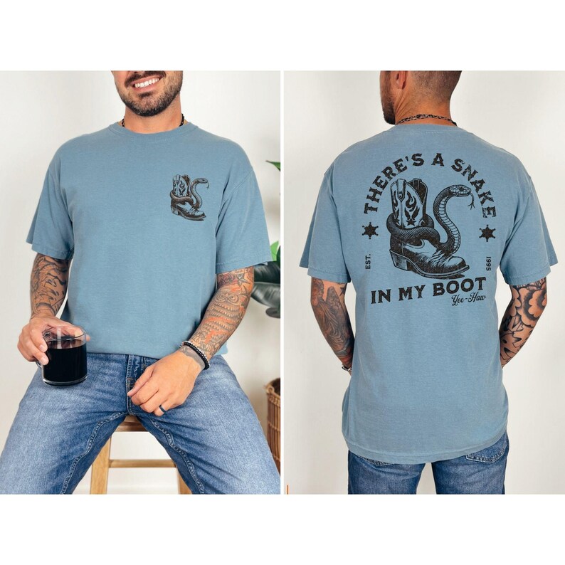 May include: Dusty blue t-shirt with a graphic of a snake coiled around a cowboy boot. The front of the shirt has a small version of the graphic. The back of the shirt has the text "THERE'S A SNAKE IN MY BOOT" with additional text and stars.