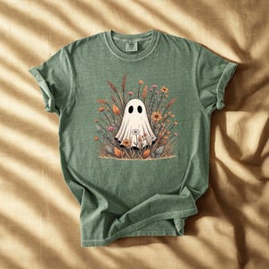 Puede incluir: Camiseta verde con una ilustración de fantasma blanco rodeado de flores y follaje coloridos. El fantasma tiene dos ojos negros. La camiseta es de material suave y tiene cuello redondo.