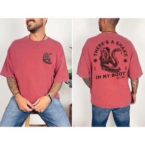 May include: A red t-shirt with a graphic of a snake coiled around a cowboy boot. The front of the shirt has a smaller version of the graphic. The back of the shirt has the text "THERE'S A SNAKE IN MY BOOT" with stars and the words "Yee-Haw".