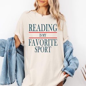 May include: Cream-colored t-shirt with the text "READING IS MY FAVORITE SPORT" in navy blue, with a red line above and below "IS MY". The shirt is a casual style.