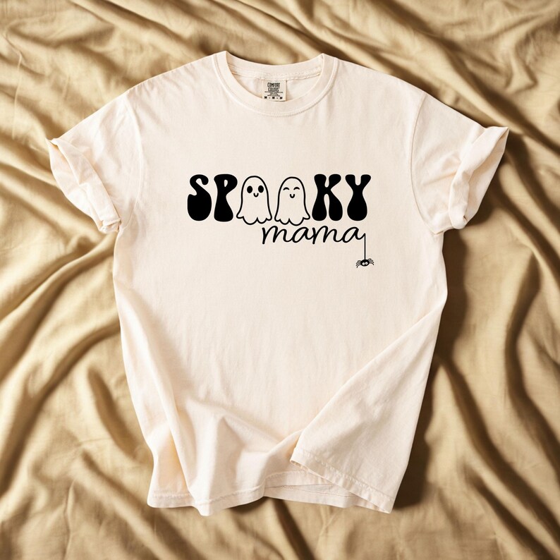 May include: Cream-colored t-shirt with the words "Spooky mama" in black, featuring two ghost illustrations and a spider. The shirt has a crew neck and short sleeves, perfect for Halloween.