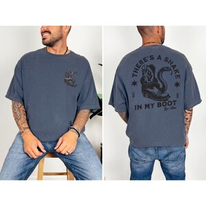 May include: A slate blue t-shirt with a graphic on the front and back. The front graphic is a snake. The back graphic says "THERE'S A SNAKE IN MY BOOT" with a snake and cowboy boot illustration. The shirt is a relaxed fit.