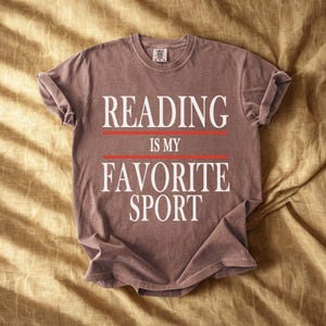 May include: A brown t-shirt with the text "READING IS MY FAVORITE SPORT" in white, with red lines separating the words. The shirt is folded and laid on a golden-yellow fabric.