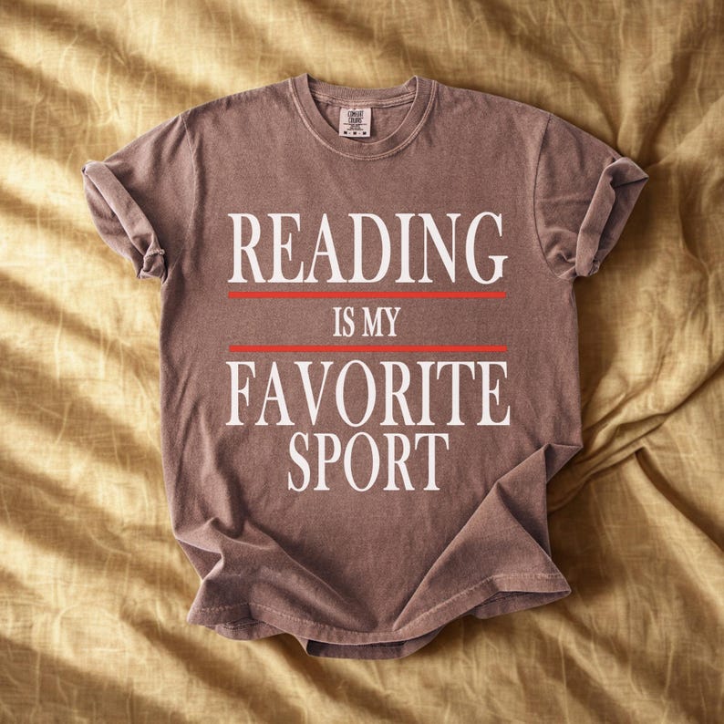 May include: A brown t-shirt with the text "READING IS MY FAVORITE SPORT" in white, with red lines separating the words. The shirt is folded and laid on a golden-yellow fabric.