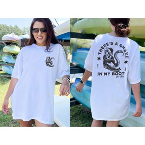 May include: White t-shirt with a graphic of a snake coiled around a cowboy boot. The text on the back of the shirt reads "THERE'S A SNAKE IN MY BOOT" with additional text and stars. The shirt is worn by a person.